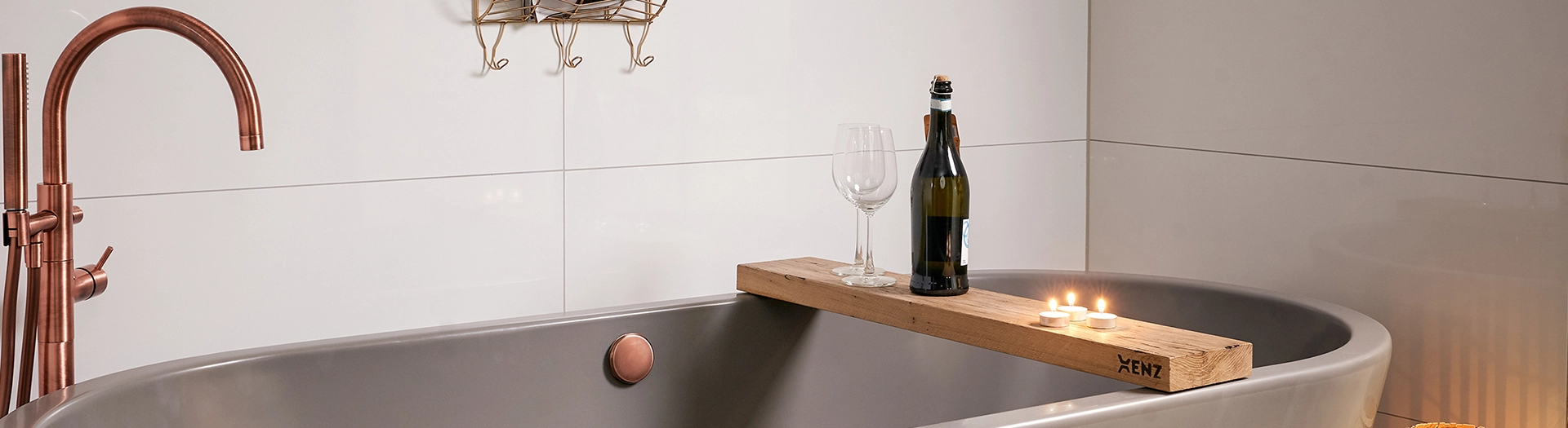 aanzicht van een koperkleurige vrijstaande badkraan, een donkergrijs vrijstaand ligbad en een eiken badplankje met kaarsjes, een fles wijn en twee wijnglazen